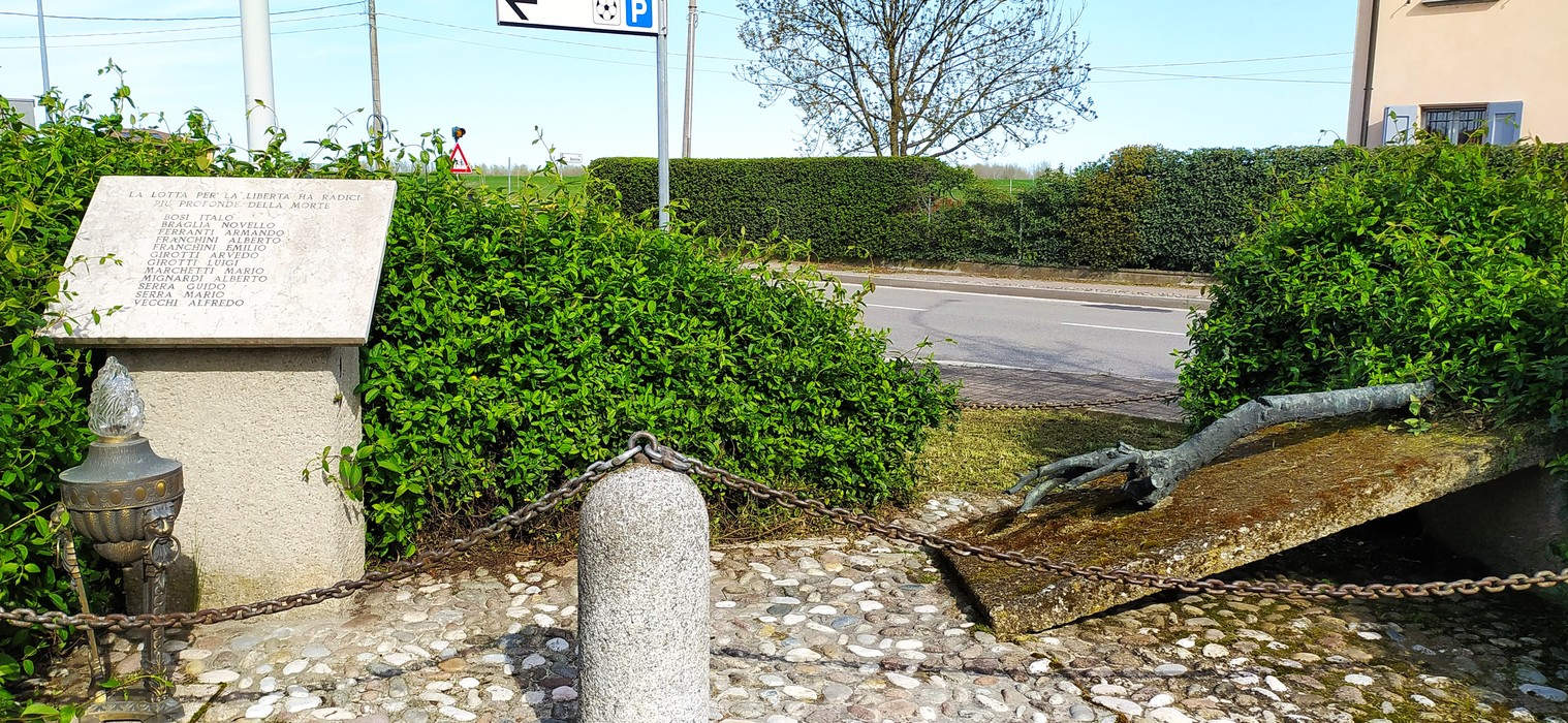 Monumento ai Caduti di Le Budrie, con la stele incorniciata da vegetazione e un braccio che emerge da una lastra.