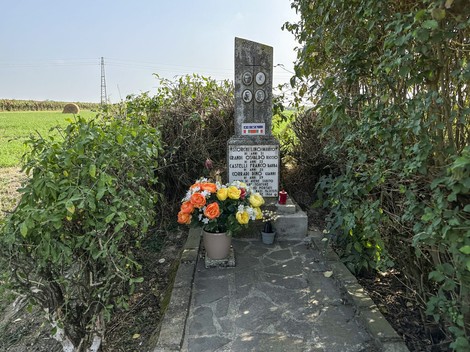 Cippo commemorativo decorato con fiori, situato in un ambiente rurale.
