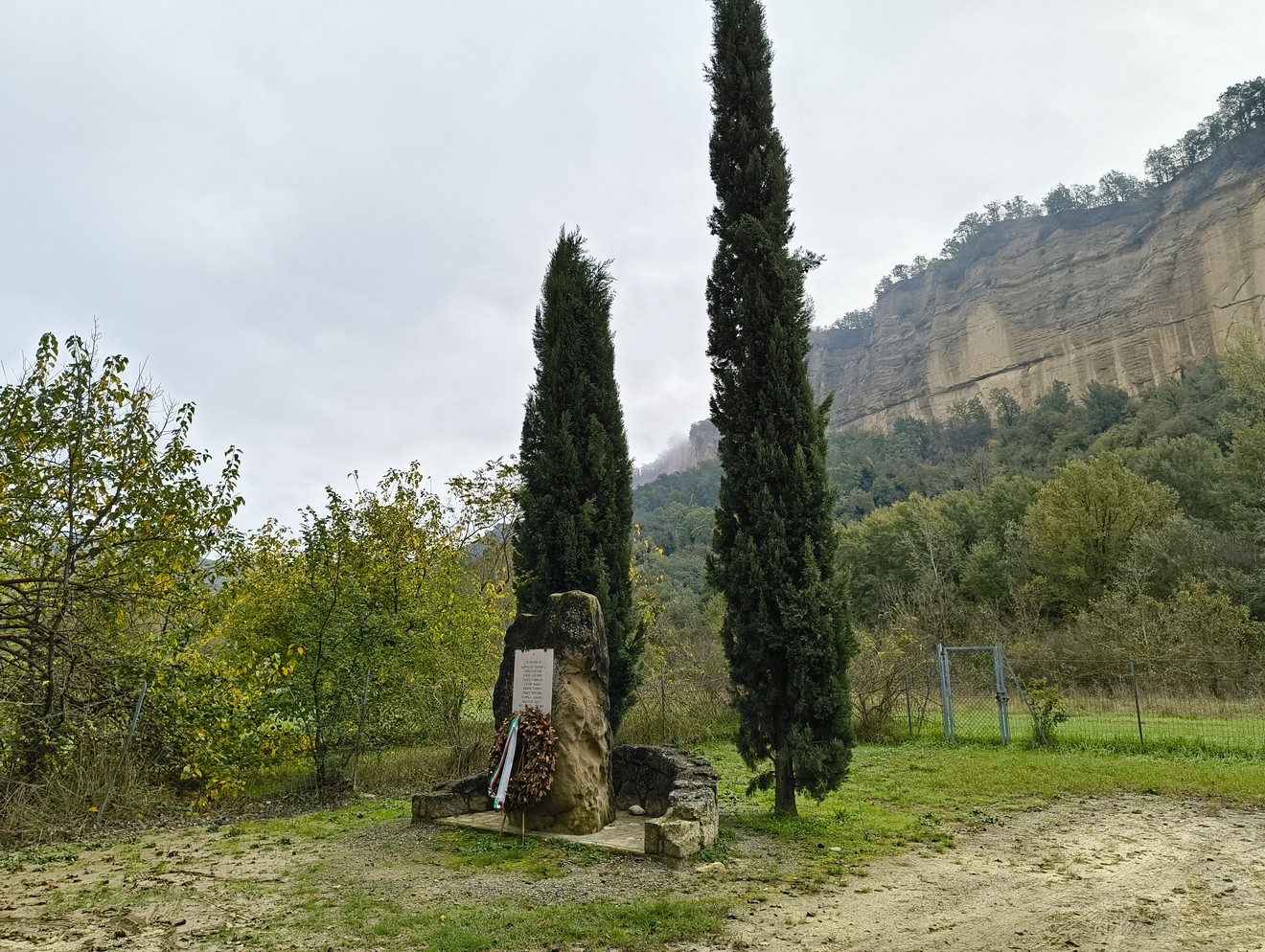 Monumento agli uccisi di Rio Conco, immerso nella natura con cipressi e scogliere sullo sfondo.