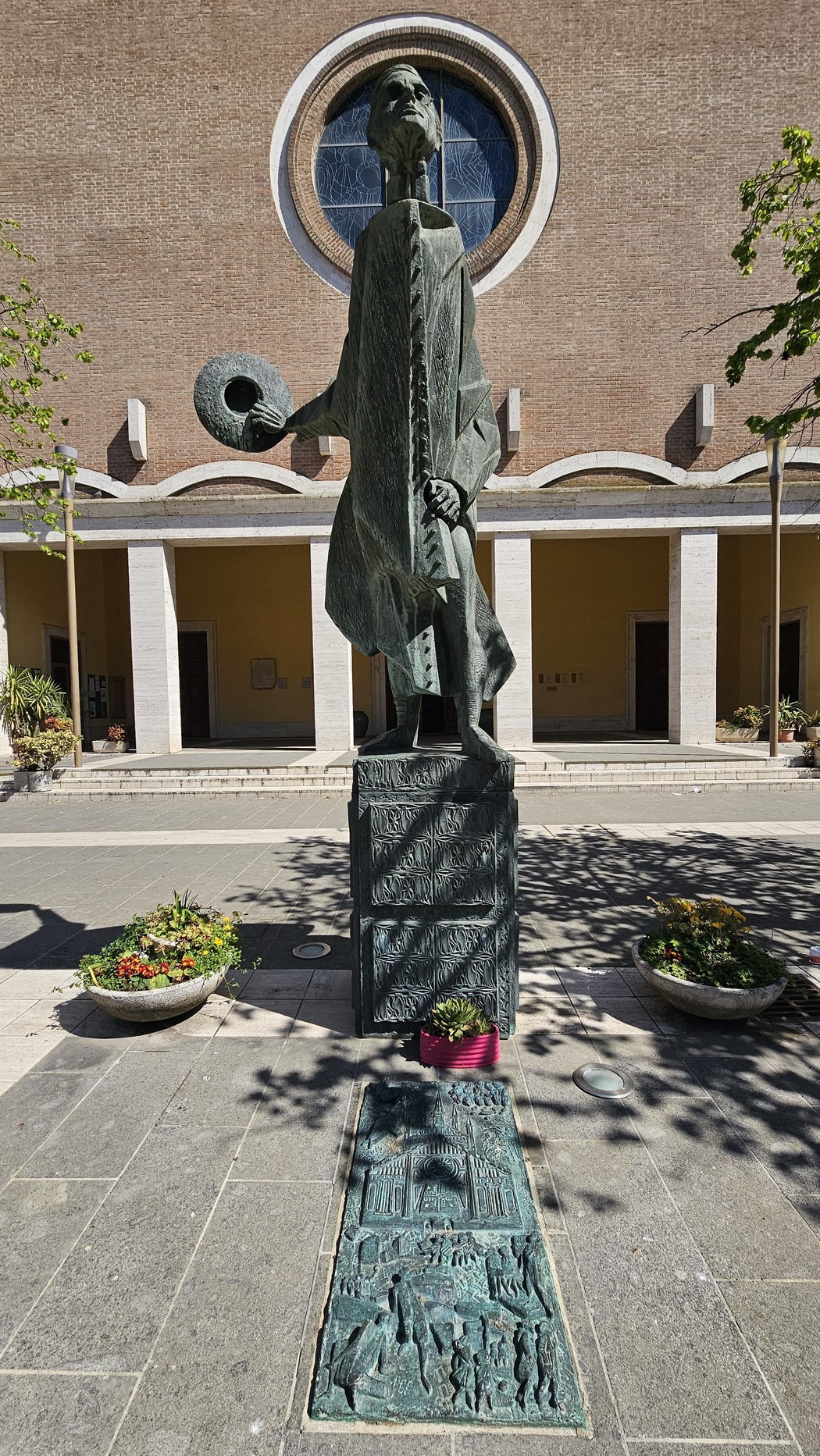 Monumento a Minzoni, una scultura con una figura maschile che tiene un cappello, circondata da fiori e architettura.