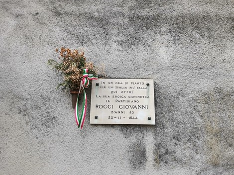 Lapide commemorativa dedicata al partigiano Giovanni Rocci, con decorazioni floreali e nastri tricolori.