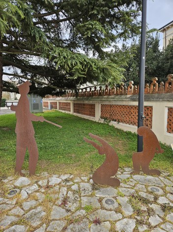 Scultura in metallo raffigurante due figure animate in Largo Martiri del Nazifascismo, circondate da verde e architettura storica.