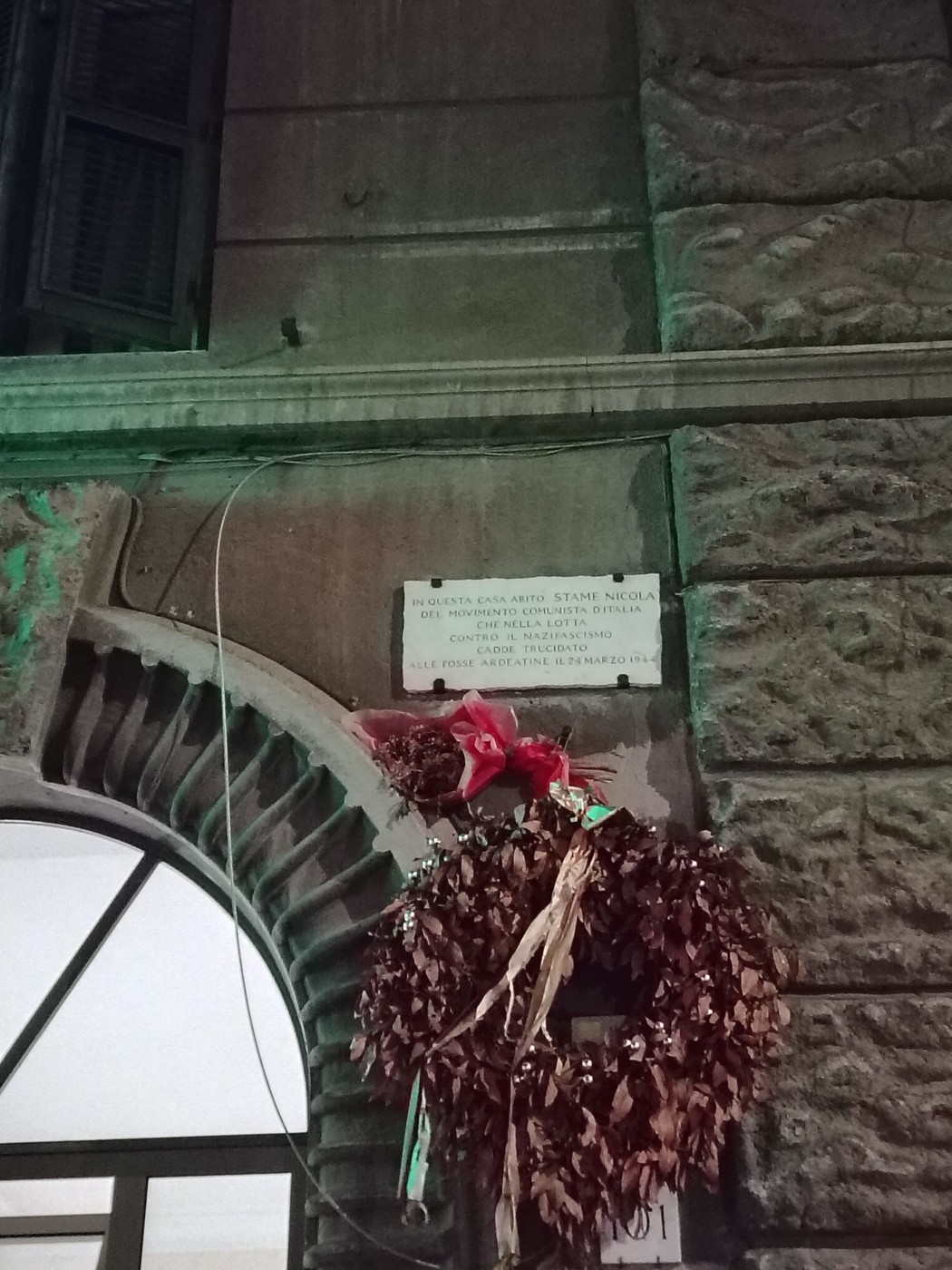 Lapide commemorativa a Stame con ghirlanda decorativa, in un contesto urbano illuminato.