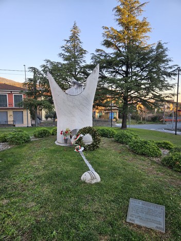 Monumento alla Resistenza circondato da verde, con decorazioni floreali e un elemento commemorativo al centro.