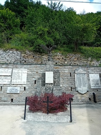 Monumento ai caduti circondato da pareti di pietra e vegetazione, con iscrizioni memoriali sui lati.