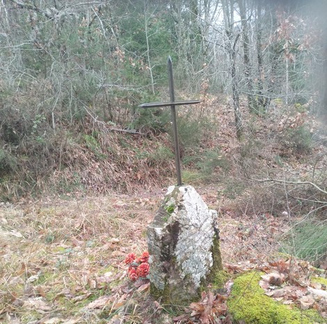 Croce metallica su un basamento di pietra, circondata da un bosco e terreno coperto di foglie.