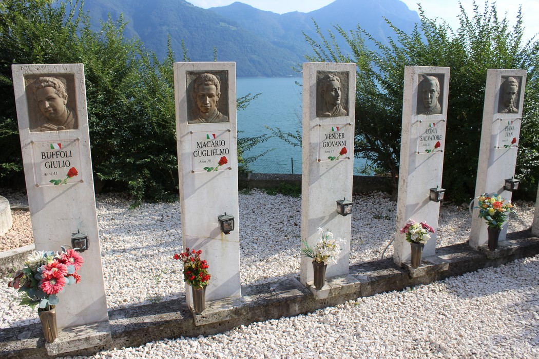 Sacrario dei Tredici Martiri, monumento commemorativo con ritratti e decorazioni floreali sullo sfondo lacustre.