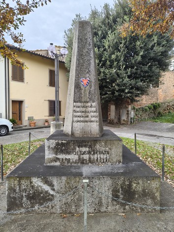 Monumento ai deportati, obelisco in pietra circondato da una catena, immerso in un contesto urbano e alberato.