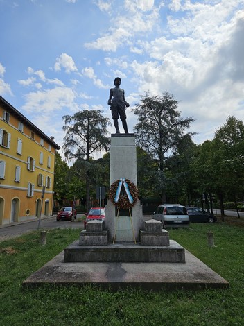 Monumento ai Caduti con una statua su un piedistallo, circondato da alberi e automobili in un contesto urbano.