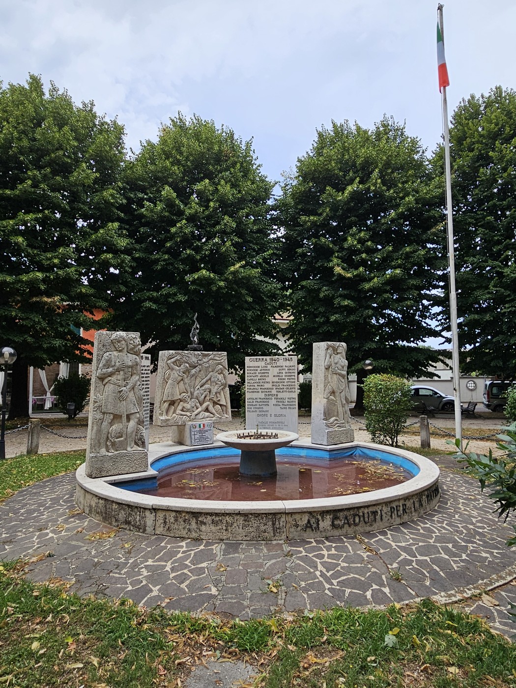 Monumento ai Caduti di San Bassano, circondato da alberi, con fontana e bandiera italiana. Un tributo ai caduti.
