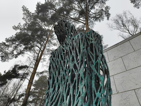 Scultura in bronzo al cimitero militare italiano di Zehlendorf, circondata da alberi. Atmosfera di riflessione.