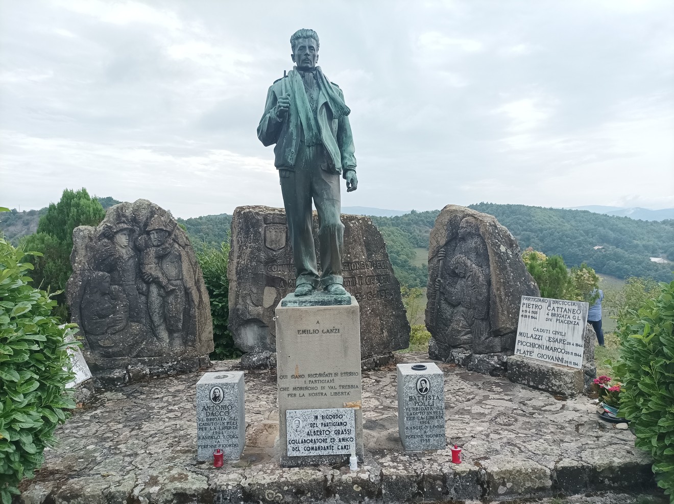 Monumento ai caduti con statua e pietre commemorative, immerso nel verde e panoramica su un paesaggio naturale.