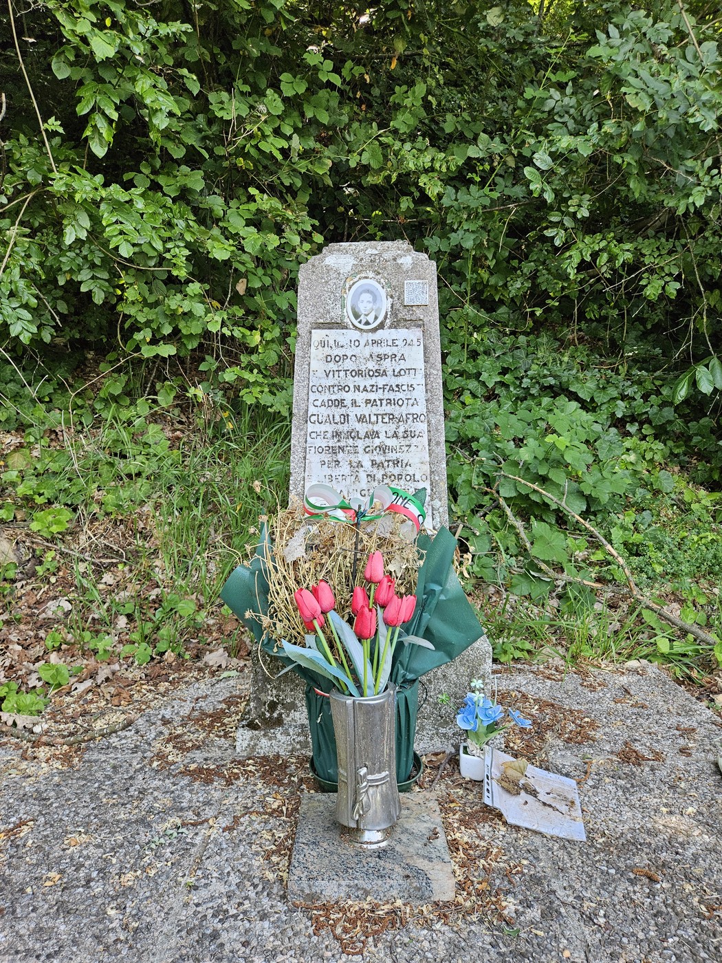 Cippo commemorativo a Gualdi, adornato con fiori e decorazioni, immerso nella vegetazione circostante.