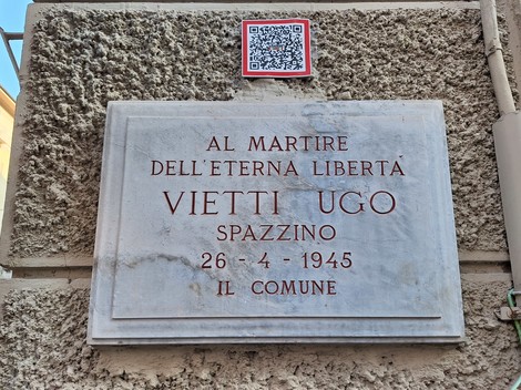 Lapide in marmo con iscrizione su parete esterna intonacata, con QR code applicato sopra.