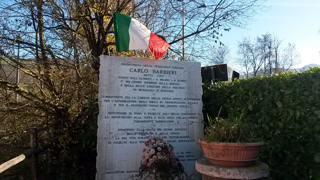 Monumento a Carlo Barbieri, con bandiera italiana, situato in un contesto naturale.