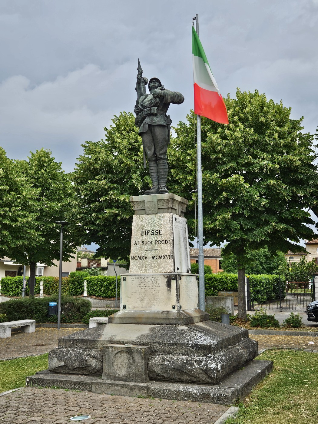 Monumento ai Caduti con statuaria commemorativa e bandiera italiana, circondato da alberi e un'area verde.