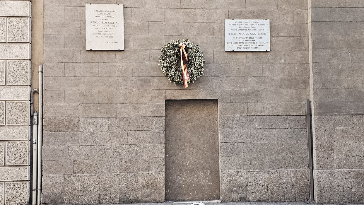 Lapide commemorativa su un muro con una corona di alloro, in ricordo di eventi storici e persone significative.