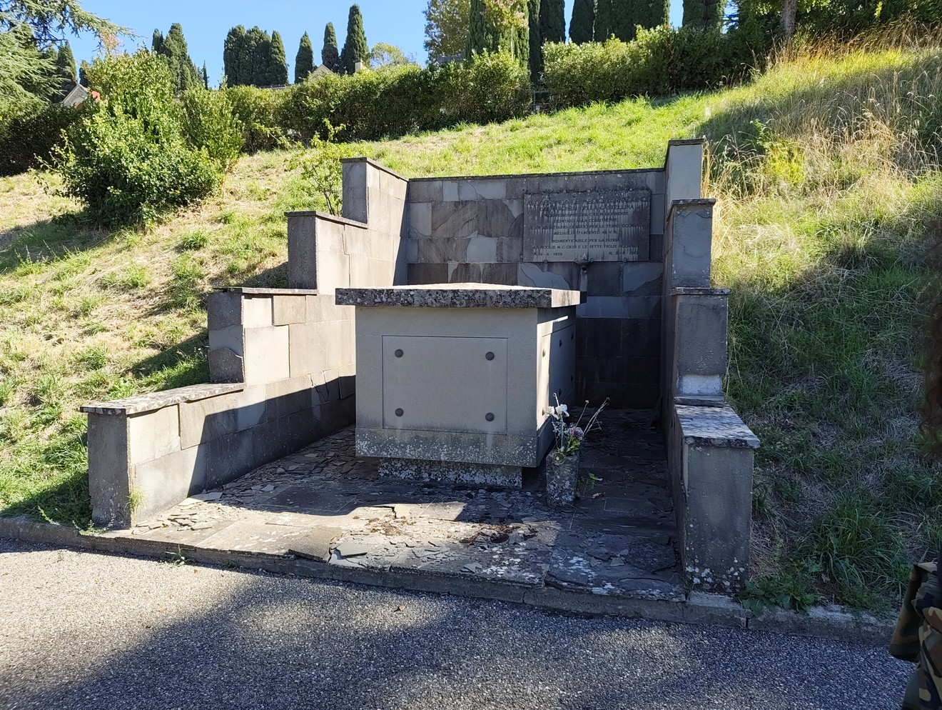 Sepolcro in pietra addossato a un pendio erboso, con lapide incisa sopra un basamento centrale.