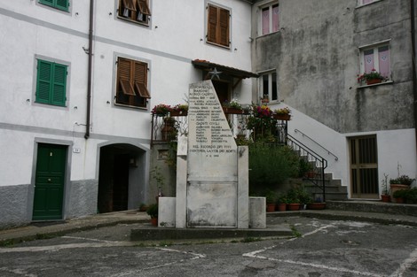 Monumento ai caduti inserito in un contesto urbano, circondato da fiori e edifici storici.