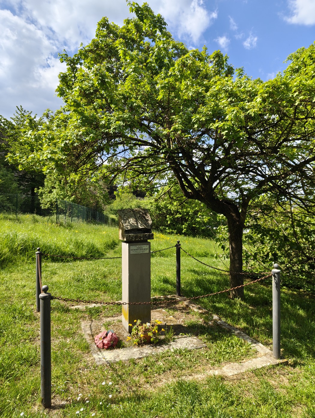 Cippo a Meuret con un albero verde e prato, circondato da una catena in un paesaggio naturale.