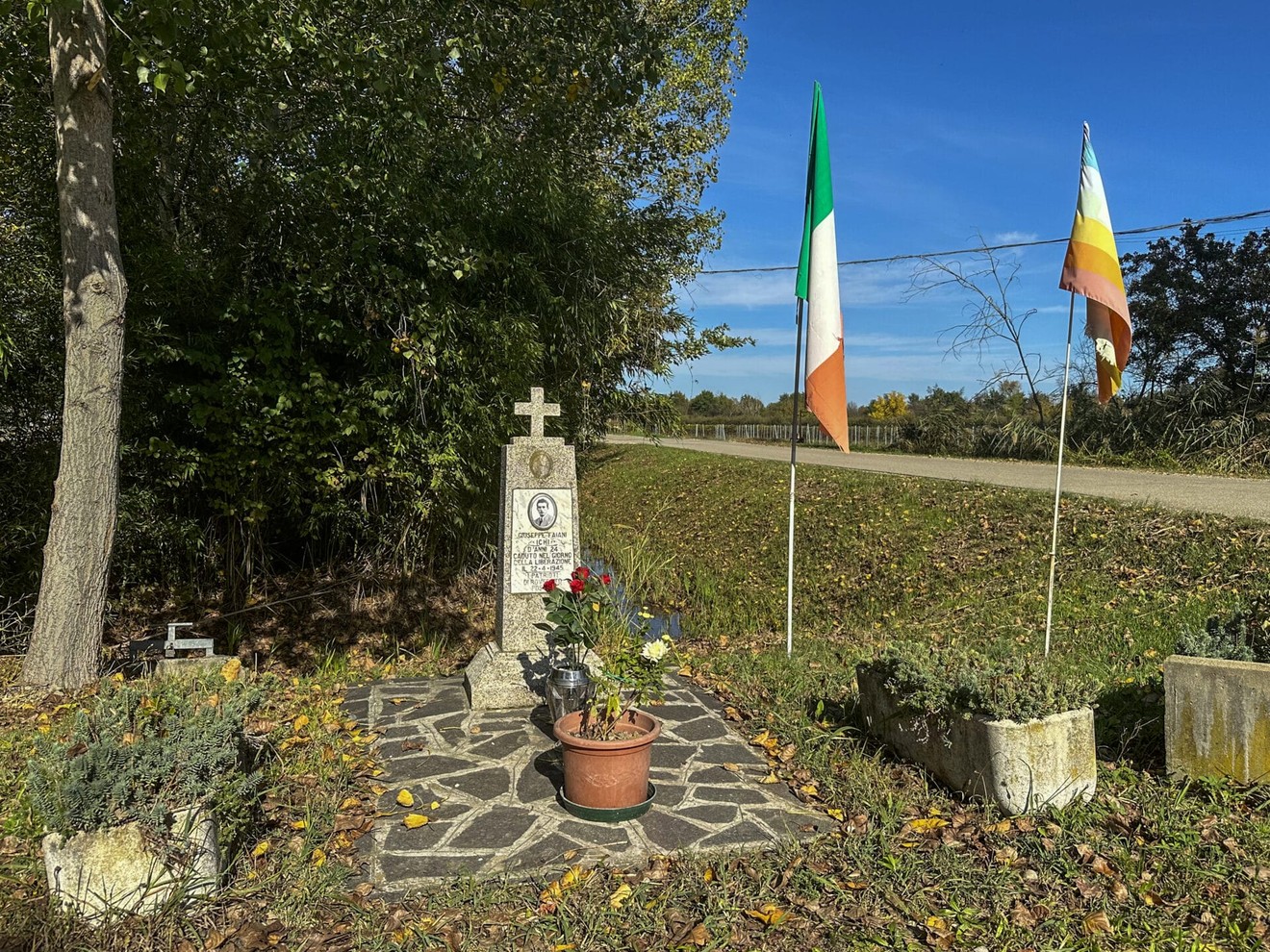 Cippo commemorativo a Faiani, adornato con fiori e bandiere, immerso nel verde di un paesaggio autunnale.