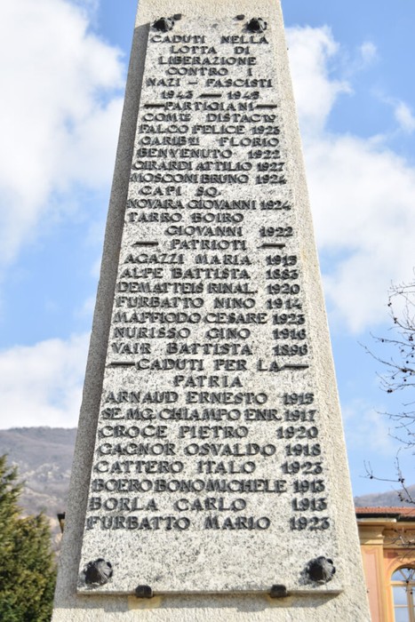 Obelisco di pietra con lunga iscrizione commemorativa, all'aperto, con alberi sullo sfondo.