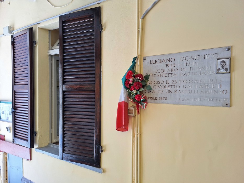 Lapide commemorativa di Luciano, con un omaggio floreale e un tricolore italiano, situata vicino a una finestra.