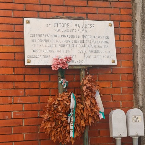 Lapide marmorea con iscrizione su parete di mattoni, sotto corone di fiori secchi appese.
