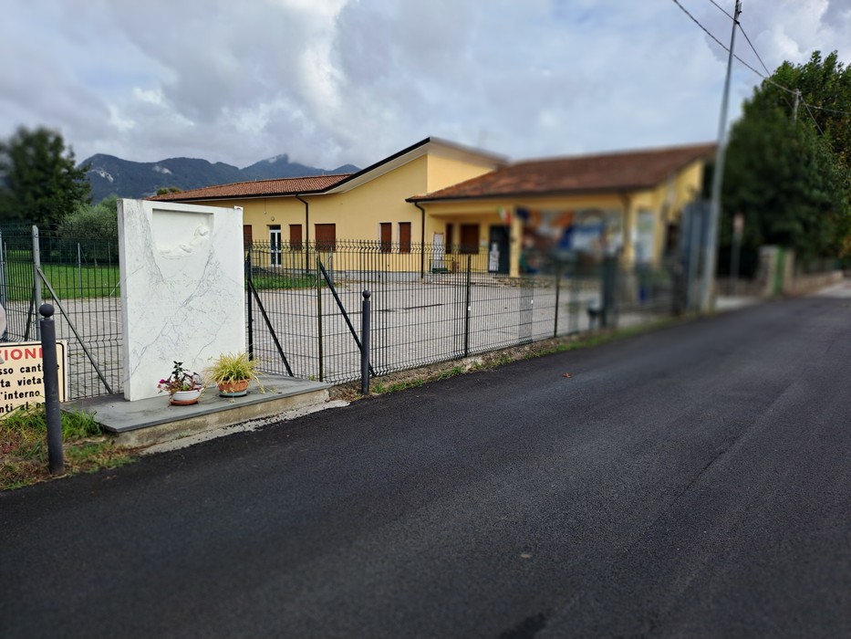 Cippo in marmo con iscrizione, recintato davanti a un edificio, con un vaso di fiori alla base.