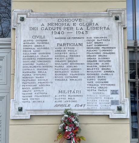 Lapide marmorea su parete esterna, con lunghi elenchi di nomi e mazzo di fiori sottostante.