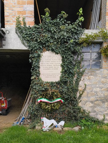 Lapide commemorativa dedicata ai martiri della Creda, circondata da vegetazione e decorazioni tricolori.