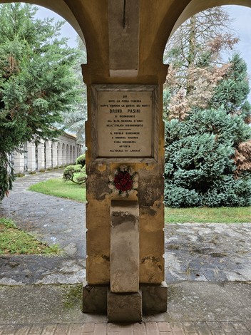 Lapide commemorativa affissa su un muro, circondata da alberi e un percorso di pietra.