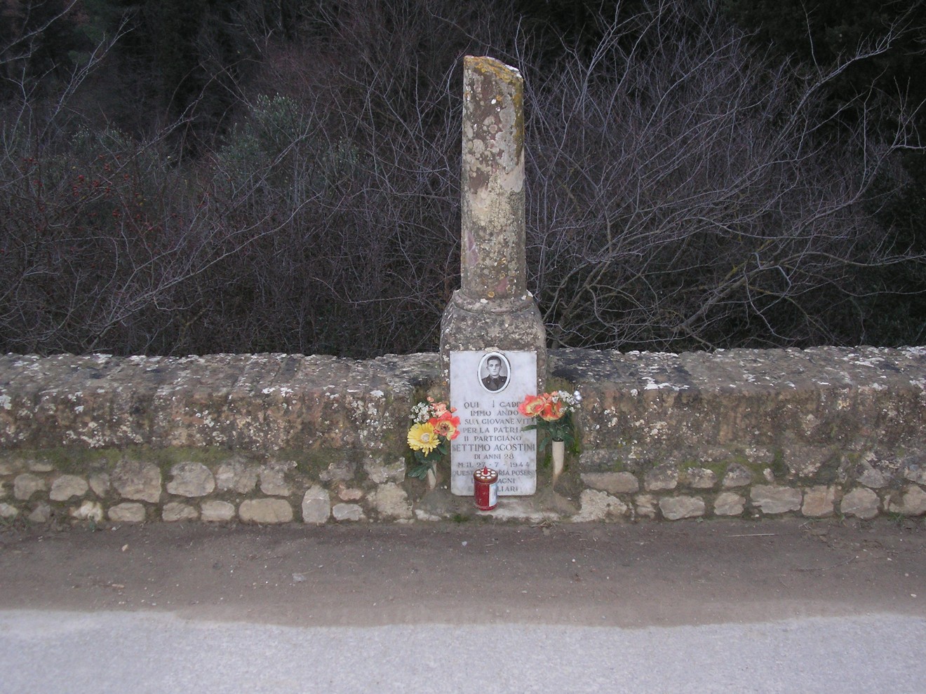 Cippo commemorativo ad Agostini, adornato con fiori e lumini, immerso nella natura circostante.