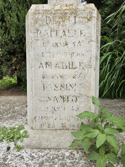 Cippo commemorativo con nomi e date di persone scomparse, circondato da vegetazione.