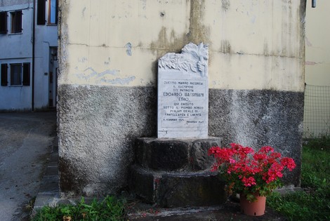 Cippo commemorativo dedicato a edoard Bassignani, con una decorazione floreale e sfondo di edifici storici.
