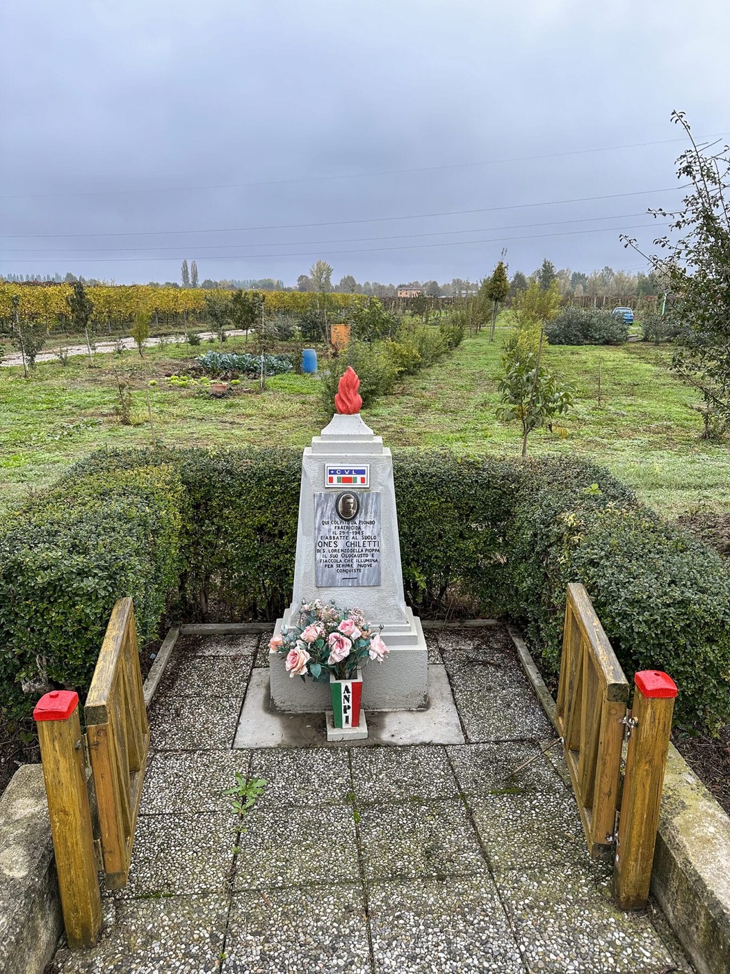Cippo commemorativo a Chiletti, circondato da vegetazione, con una decorazione floreale e bandiera italiana.