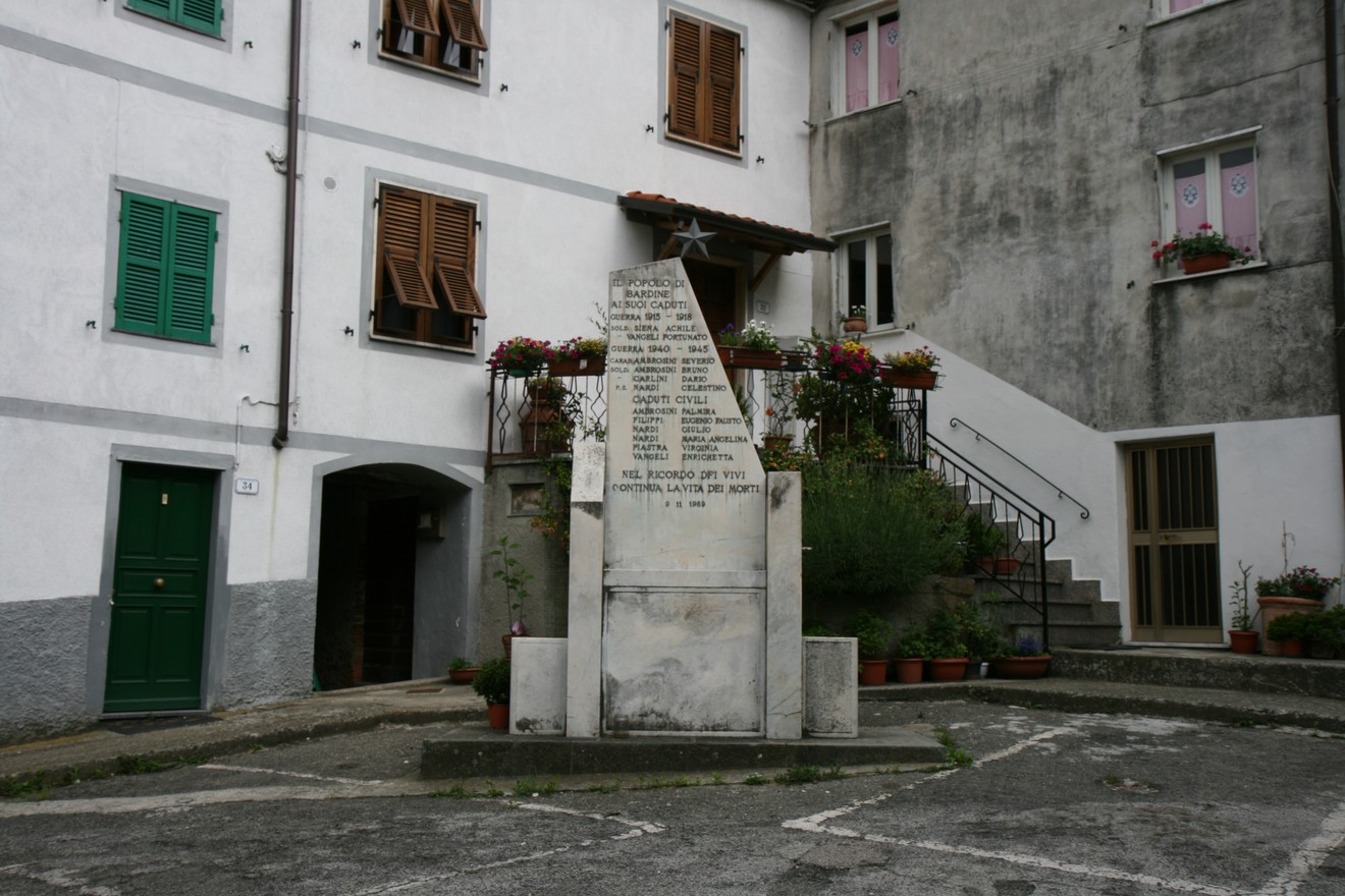 Monumento ai caduti inserito in un contesto urbano, circondato da fiori e edifici storici.
