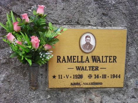 Targa commemorativa dedicata a Walter Ramella, con fiori artificiali e immagine in un contesto di memoria storica.