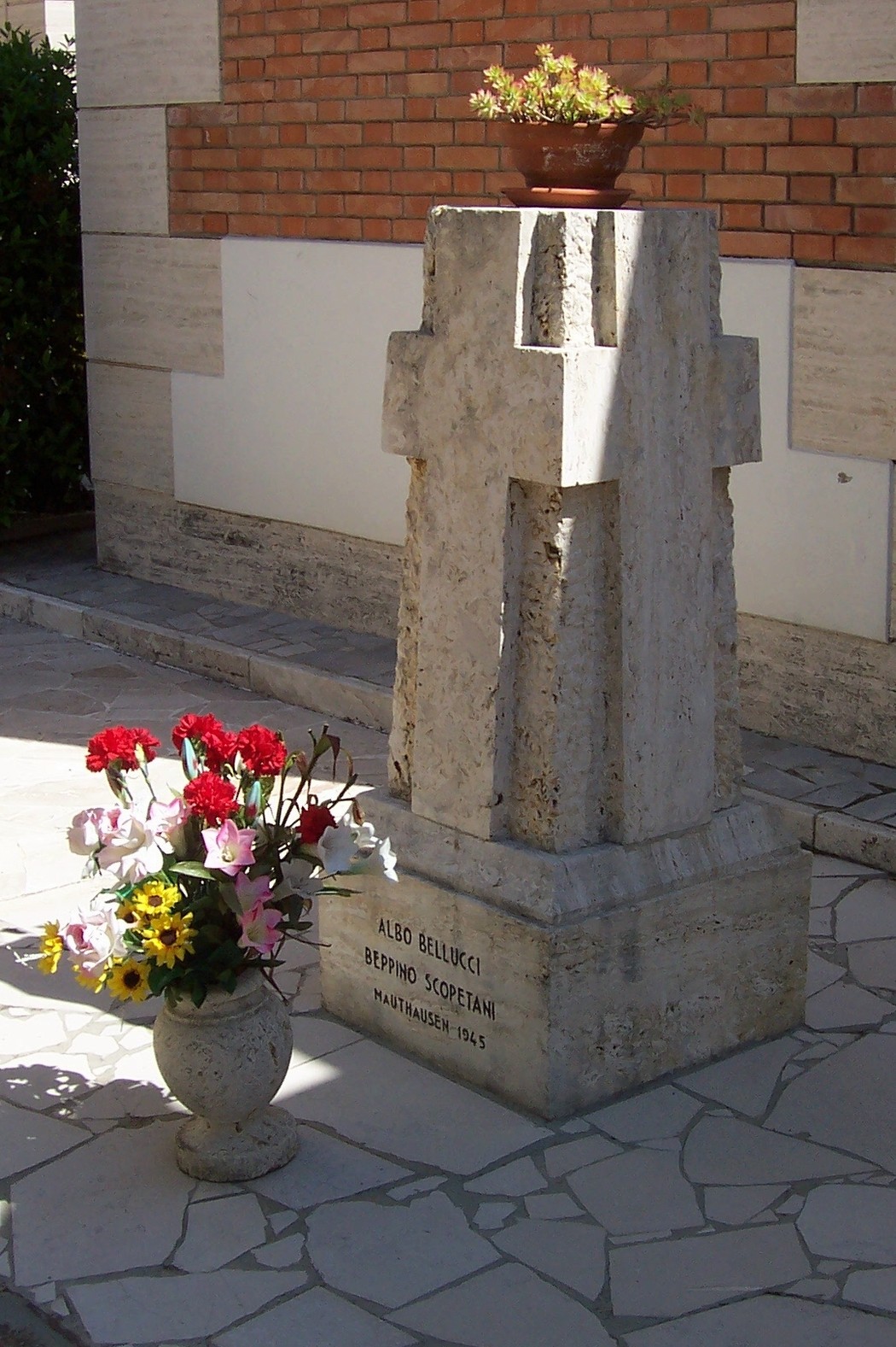 Cippo commemorativo dedicato a Bellucci e Scopetani, adornato da fiori e piante in un contesto tranquillo.