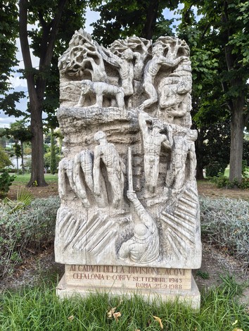 Monumento in pietra scolpita con figure in rilievo e iscrizione, collocato in un’area alberata.