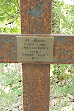Croce arrugginita con targa commemorativa in un contesto naturalistico.