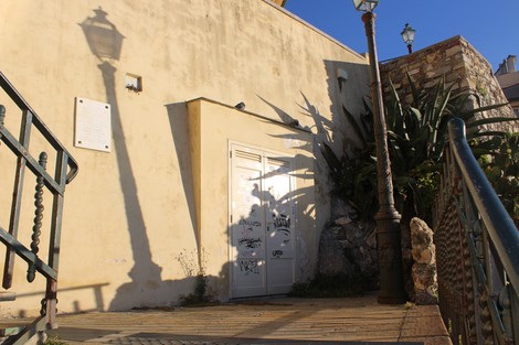 Lapide ai deportati su una parete, accanto a una porta e illuminata da un lampione, con piante e ombre evidenti.