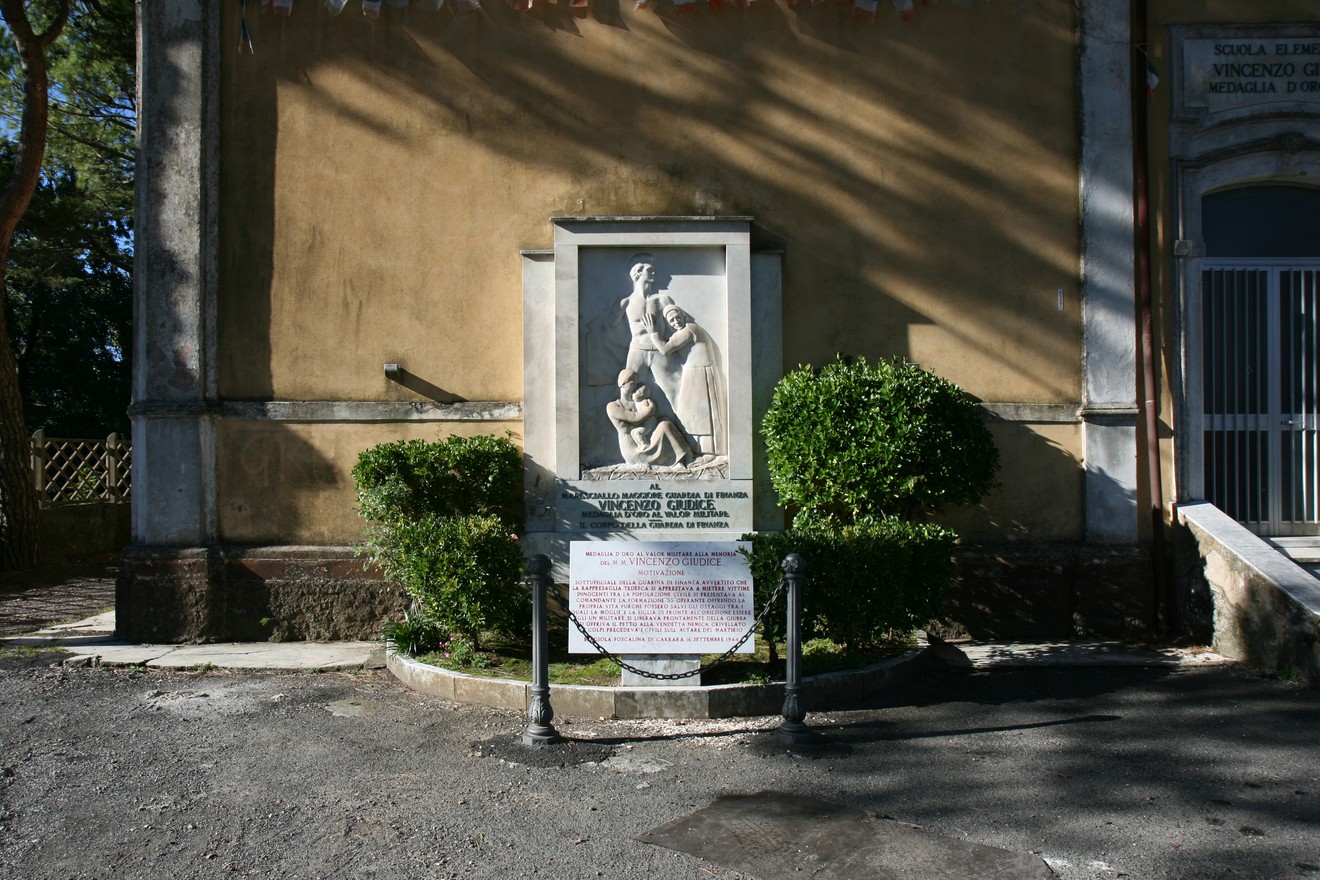Monumento a Giudice, scultura commemorativa situata vicino a un edificio storico, circondata da verde.