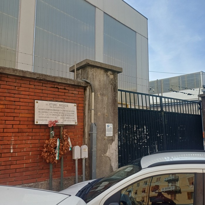 Lapide in marmo su muro di mattoni, con fiori deposti sotto, accanto a un cancello.