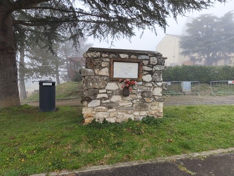 Lapide commemorativa in pietra, circondata da vegetazione e nebbia, con fiori freschi.