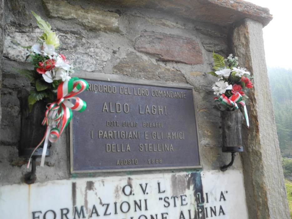 Targa in metallo su parete in pietra, con iscrizione, decorata da mazzi di fiori ai lati.