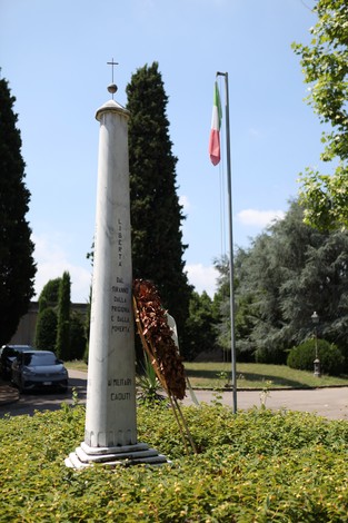 Monumento ai caduti con obelisco bianco, corona di alloro e bandiera italiana sullo sfondo.