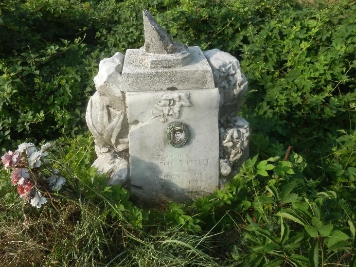 Cippo commemorativo immerso nella vegetazione, con decorazioni floreali e un elemento appuntito sulla sommità.