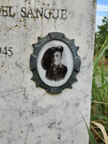 Cippo funerario con fotografia di un uomo in uniforme, circondato da vegetazione in un contesto rurale.