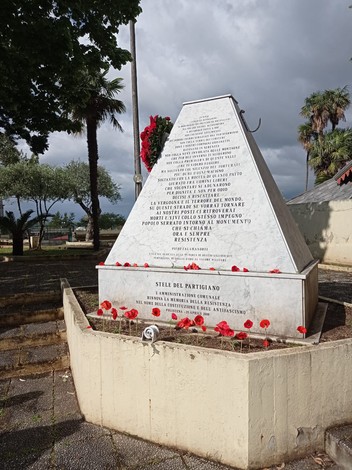 Monumento dedicato ai partigiani, con iscrizioni memoriali e fiori rossi, incorniciato da un paesaggio naturale.